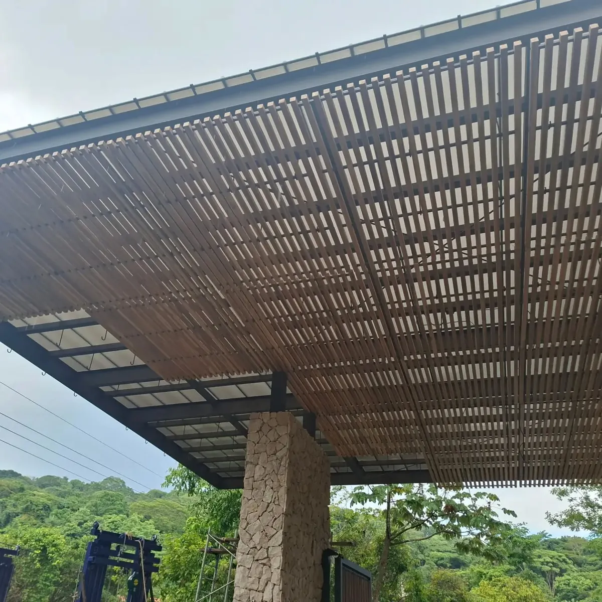 Vista del pórtico durante la instalación, con paneles de teca sobre estructura de hierro y columna de piedra